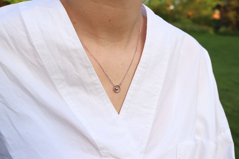 May include: A silver necklace with a small pendant featuring a sphere within a circle. The necklace is worn over a white shirt with a V-neck. The background is blurred, suggesting an outdoor setting.