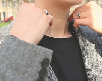May include: A person wearing a gray herringbone blazer over a black t-shirt and a silver chain necklace. The person is wearing a silver ring on their left hand and two silver rings on their right hand.