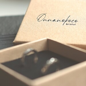 May include: A small, brown cardboard box with the text "Onnanofoco Bristol" printed on the lid. The box is open and two silver rings are visible inside.