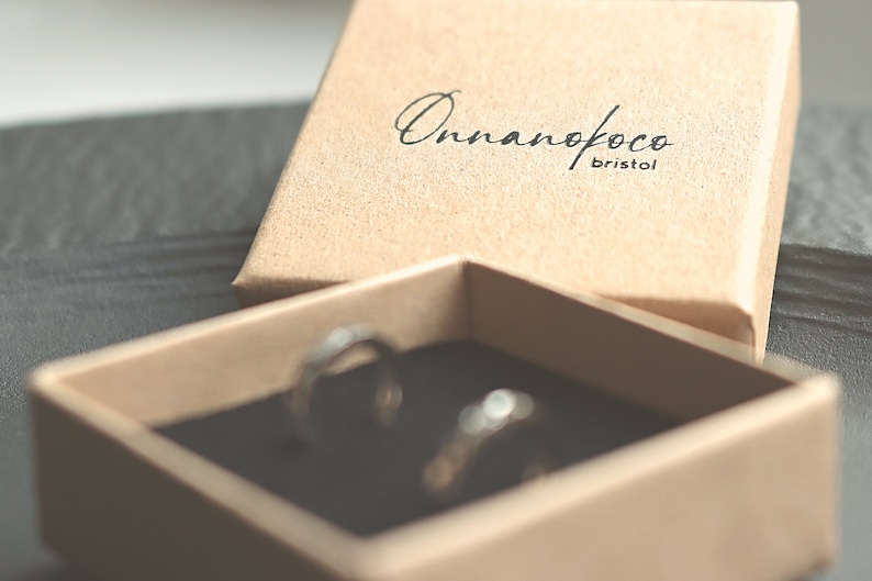 May include: A small, brown cardboard box with the text "Onnanofoco Bristol" printed on the lid. The box is open and two silver rings are visible inside.