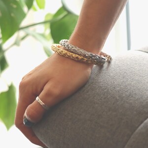 May include: Two braided bracelets, one gold and one silver, worn on a person's wrist.