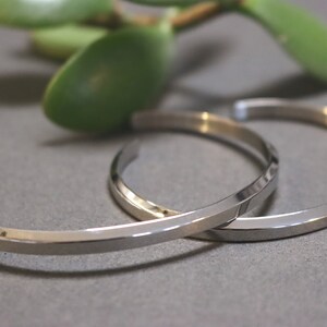 May include: Two silver metal bracelets with a flat, angular design. The bracelets are stacked on top of each other.