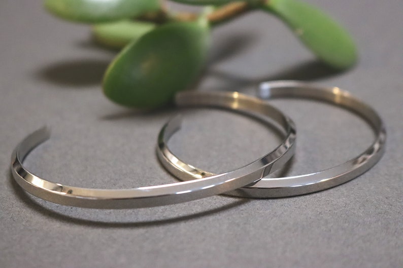 May include: Two silver metal bracelets with a flat, angular design. The bracelets are stacked on top of each other.