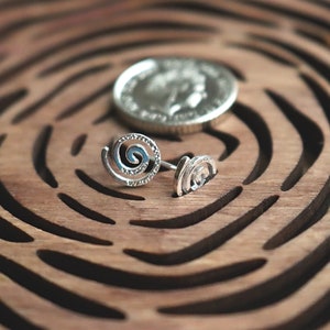 May include: Two silver spiral earrings on a wooden surface with a carved pattern. The earrings are small and delicate, with a simple design.