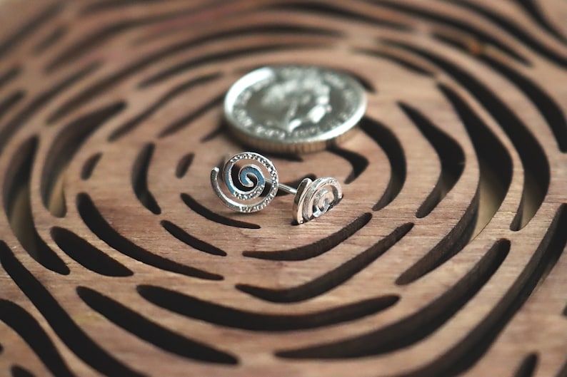 May include: Two silver spiral earrings on a wooden surface with a carved pattern. The earrings are small and delicate, with a simple design.