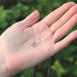 May include: A delicate silver necklace featuring a small, round pendant within an oval frame. The pendant has a small, round, shiny accent. The necklace is displayed against a blurred green background.