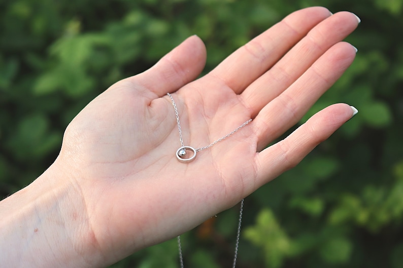 May include: A delicate silver necklace featuring a small, round pendant within an oval frame. The pendant has a small, round, shiny accent. The necklace is displayed against a blurred green background.