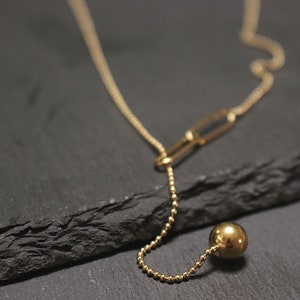 May include: A gold necklace with a long, thin chain and a single, large, round pendant. The necklace is on a black surface.