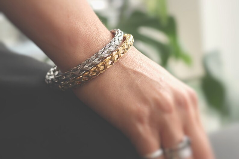 May include: Two braided bracelets, one gold and one silver, worn on a person's wrist.