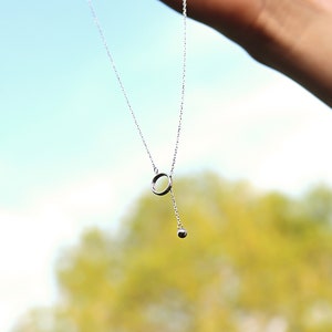 May include: A delicate silver necklace with a circular pendant and a small dangling sphere. The necklace is held against a backdrop of a blurred green and blue sky, suggesting an outdoor setting. The jewelry is simple and elegant.