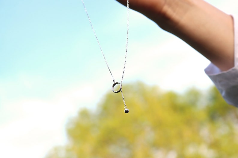 May include: A delicate silver necklace with a circular pendant and a small dangling sphere. The necklace is held against a backdrop of a blurred green and blue sky, suggesting an outdoor setting. The jewelry is simple and elegant.