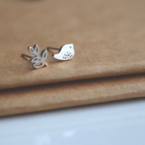 Może przedstawiać: Para srebrnych kolczyków, jeden z małym wzorem ptaka, a drugi z prostym wzorem liścia.