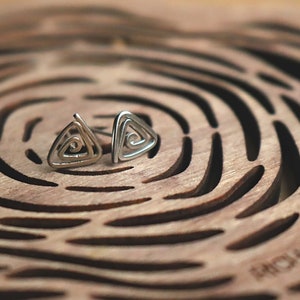May include: A pair of silver triangle earrings with a spiral design. The earrings are on a wooden surface with a circular pattern.