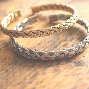 May include: Two braided metal bracelets, one gold and one silver, on a wooden surface.