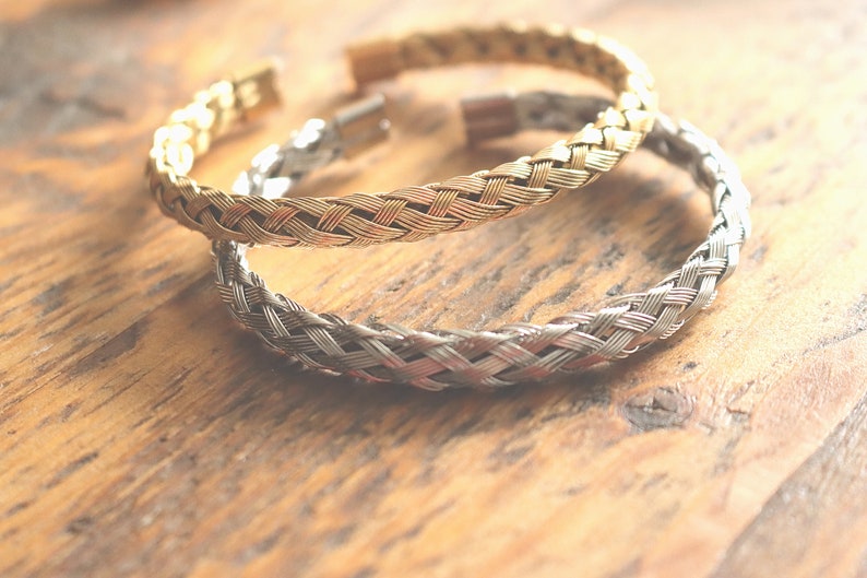 May include: Two braided metal bracelets, one gold and one silver, on a wooden surface.