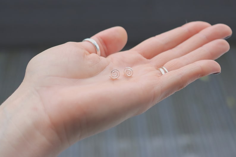 May include: A pair of silver spiral stud earrings on a hand.