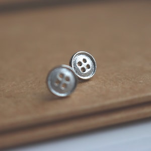 May include: A pair of silver button earrings with four holes each. The earrings are on a brown surface.