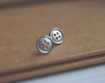 May include: A pair of silver button earrings with four holes each. The earrings are on a brown surface.
