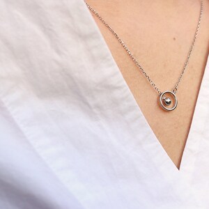 May include: A delicate silver necklace featuring a small sphere pendant within a circular frame. The necklace hangs on a person's neck, against a white shirt. The minimalist design offers a touch of elegance.