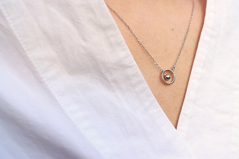 May include: A delicate silver necklace featuring a small sphere pendant within a circular frame. The necklace hangs on a person's neck, against a white shirt. The minimalist design offers a touch of elegance.