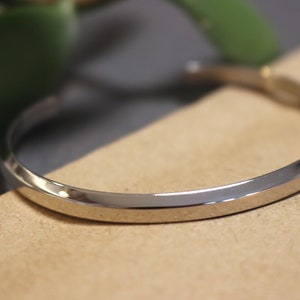 May include: A silver bangle bracelet with a smooth, rounded surface. The bracelet is made of metal and has a simple, elegant design.