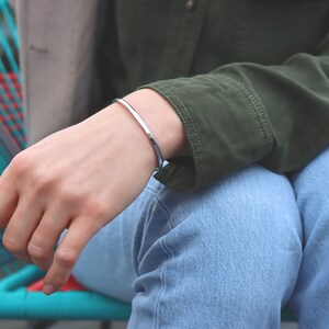 May include: A silver bangle bracelet on a person's wrist. The bracelet is simple and sleek, with a polished finish.
