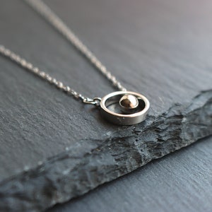 May include: A silver-toned necklace featuring a small sphere within a circular pendant. The pendant hangs from a delicate chain. The jewelry is displayed on a dark, textured surface, creating a contrast that highlights the necklace's design.
