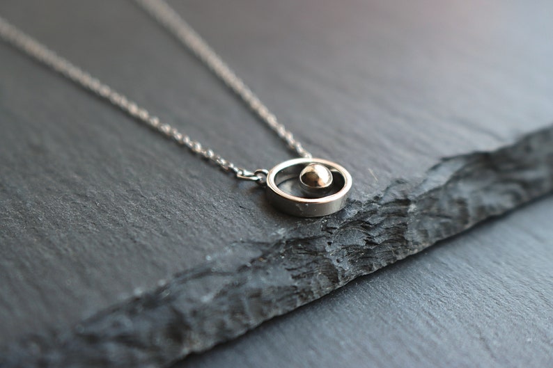 May include: A silver-toned necklace featuring a small sphere within a circular pendant. The pendant hangs from a delicate chain. The jewelry is displayed on a dark, textured surface, creating a contrast that highlights the necklace's design.