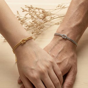 Puede incluir: Dos pulseras, una dorada y otra plateada, cada una con un diseño de nudo. La pulsera dorada está en una muñeca con una delicada cadena. La pulsera plateada está en otra muñeca. El fondo es una superficie de madera clara.