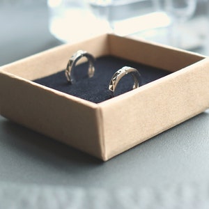 May include: Two silver hoop earrings with a geometric design are displayed in a brown box with a black velvet lining.
