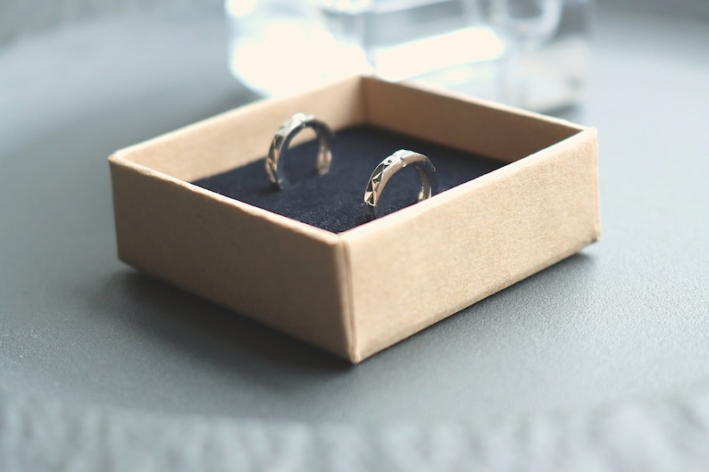 May include: Two silver hoop earrings with a geometric design are displayed in a brown box with a black velvet lining.