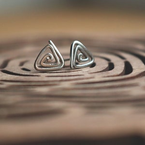 Op de afbeelding: Een paar zilveren driehoekige oorbellen met een spiraalvormig ontwerp. De oorbellen liggen op een houten oppervlak met een cirkelvormig patroon.