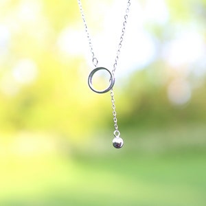 May include: A delicate silver necklace featuring a small open circle pendant connected to a chain. A dangling silver ball hangs below the circle. The background is a soft, blurred green and yellow.