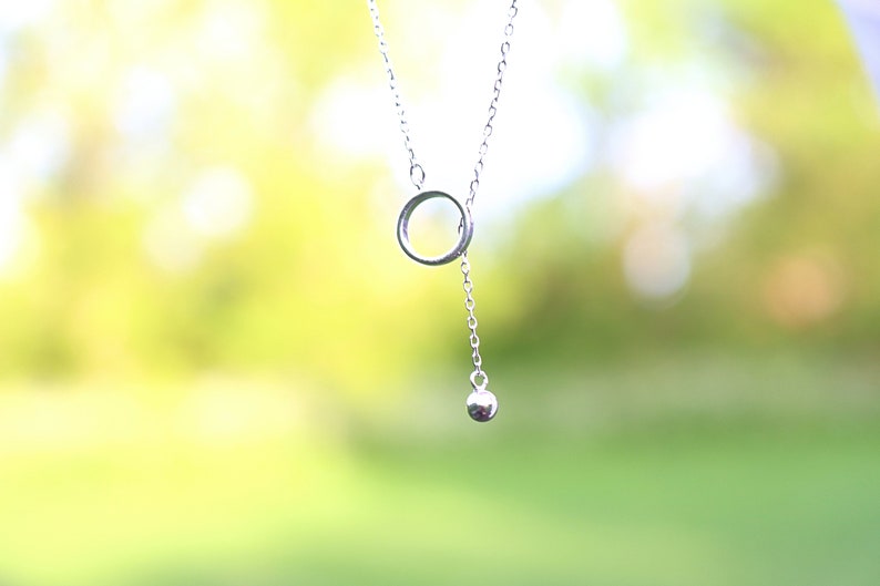 May include: A delicate silver necklace featuring a small open circle pendant connected to a chain. A dangling silver ball hangs below the circle. The background is a soft, blurred green and yellow.