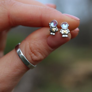 May include: A pair of silver astronaut stud earrings. Each earring features a small astronaut figure with a helmet and a star on the chest. The helmets have an iridescent, colorful sheen.