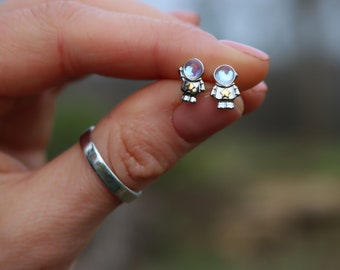 May include: A pair of silver astronaut stud earrings. Each earring features a small astronaut figure with a helmet and a star on the chest. The helmets have an iridescent, colorful sheen.