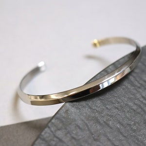 May include: A silver bangle bracelet with a slightly angled, flat surface. The bracelet is open and has a small gold accent at the clasp.