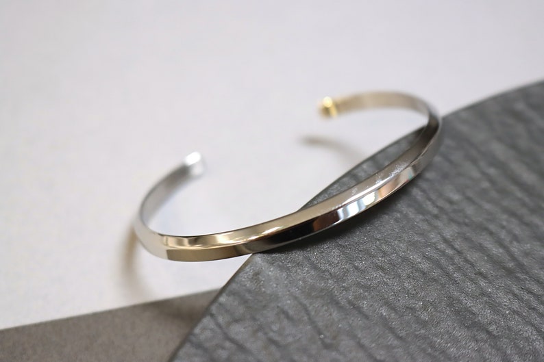 May include: A silver bangle bracelet with a slightly angled, flat surface. The bracelet is open and has a small gold accent at the clasp.