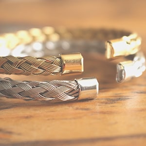 May include: A braided metal bracelet with a gold and silver finish. The bracelet is open and has a clasp on one end.