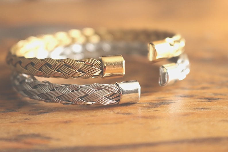 May include: A braided metal bracelet with a gold and silver finish. The bracelet is open and has a clasp on one end.