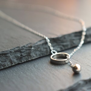 May include: A silver necklace featuring a delicate chain, a small ring pendant, and a dangling sphere. The jewelry is displayed on a dark, textured surface, highlighting the minimalist design and metallic shine.