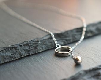 May include: A silver necklace featuring a delicate chain, a small ring pendant, and a dangling sphere. The jewelry is displayed on a dark, textured surface, highlighting the minimalist design and metallic shine.