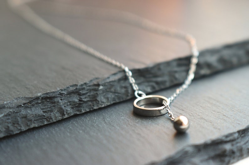 May include: A silver necklace featuring a delicate chain, a small ring pendant, and a dangling sphere. The jewelry is displayed on a dark, textured surface, highlighting the minimalist design and metallic shine.