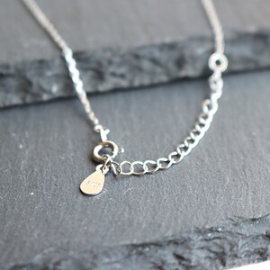 May include: Close-up of a silver necklace with a delicate chain and a small teardrop-shaped tag. The necklace features a lobster clasp closure and an adjustable chain section. The tag is stamped with the text "925".