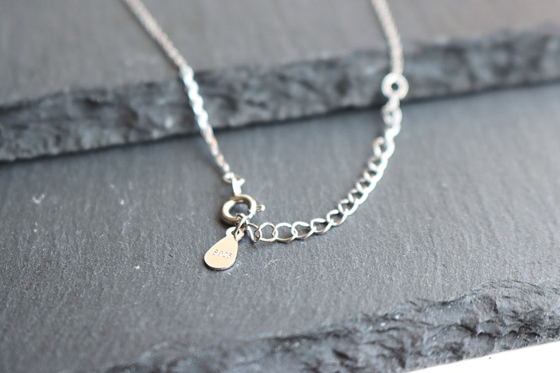May include: Close-up of a silver necklace with a delicate chain and a small teardrop-shaped tag. The necklace features a lobster clasp closure and an adjustable chain section. The tag is stamped with the text "925".