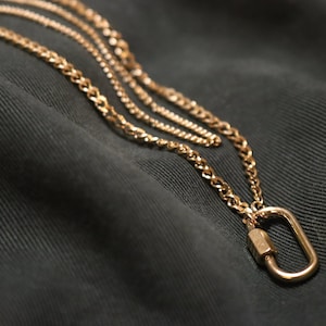May include: A gold chain necklace with a gold oval-shaped clasp. The necklace is layered on a dark fabric background.