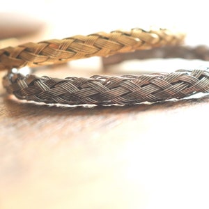 May include: Two braided metal bracelets, one gold and one silver, on a wooden surface.