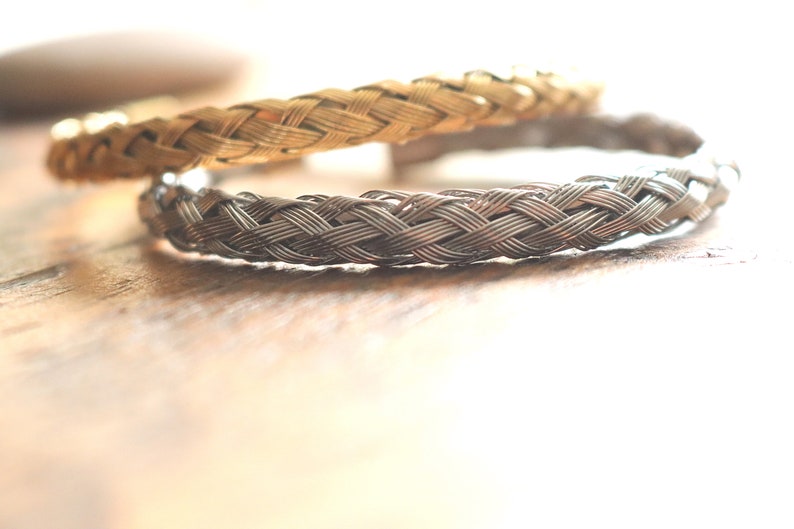 May include: Two braided metal bracelets, one gold and one silver, on a wooden surface.