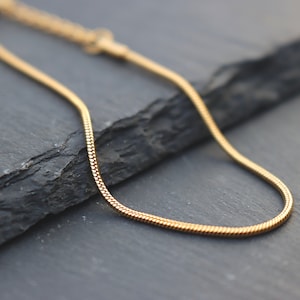 Puede incluir: Un collar de cadena de oro con un diseño de cadena de serpiente. La cadena está sobre una superficie gris oscuro.