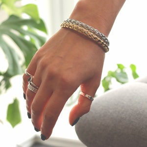 May include: A hand wearing a braided silver and gold bracelet and several silver rings.